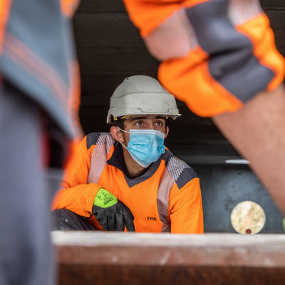 En person i hjälm och annan utrustning på ett varv jobbar. Han bär munskydd.