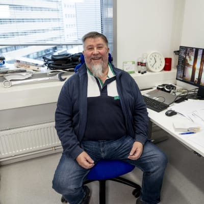 Farkkuihin ja siniseen paitaa pukeutunut Arto Orpana istuu toimistotuolilla ja hymyilee nauraen kameralle. Vieressä papereiden täyttämä työpöytä, jossa kaksi näyttöä. Näyttöjen taustakuvissa aurinko laskee vesirajaan puiden runkojen lomasta. 