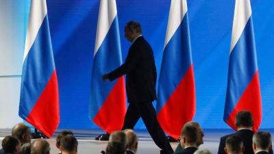 I bakgrunden flera ryska flaggor, framför den en kostymklädd man som går och i förgrunden huvuden tillhörande en publik.  Rysslands president Vladimir Putin lämnar podiet efter sitt tal till nationen den 15 januari 2020.