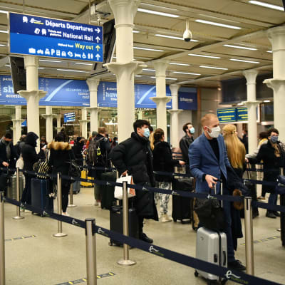 Människor köar i en tågterminal. De bär munskydd och har resväskor i händerna.