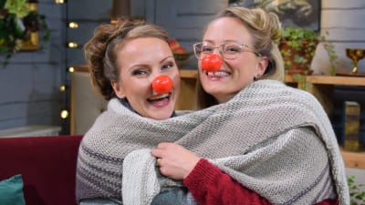 Två kvinnor med röda clownnäsor som skrattar och kramas och har lindat en stickad sjal runt sig.