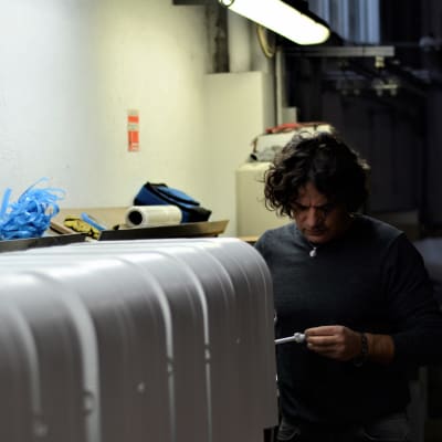 En man med halvlångt hår och grå tröja tittar fundersamt på en komponent i sin hand stående i sin fabrik.
