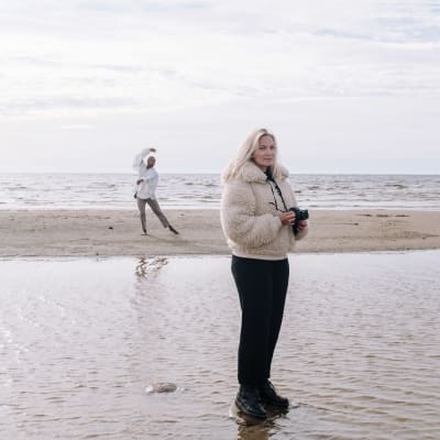 Emilia Hernesniemi ja Jani Toivola Hailuodossa meren rannalla. Toivola tanssii taustalla valkoisessa paidassa ja Hernesniemi seisoo etualalla kamera käsissään.