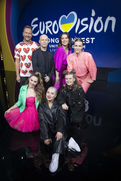 En gruppbild på sju personer i en tv-studio, tre personer sitter på golvet och fyra personer står bakom dem. I bakgrunden logon för Eurovision Song Contest 2023.