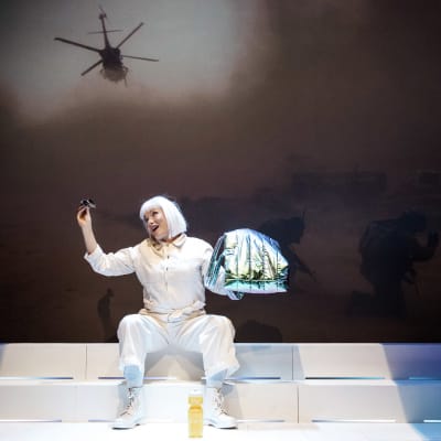 En skådespelare i vitt (Hellen Willberg) leker med ett par solglasögon medan en stridshelikopter och soldater projiceras på väggen bakom henne. 