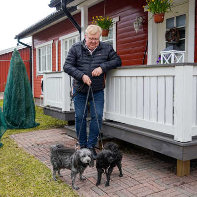 En man i jeans och svart jacka lutar sig mot ett räcke invid ett rött hus. Han håller i kopplet till två små lurviga hundar och tittar leende ner mot dem.