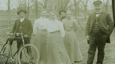 Gruppbild med fem ungdomar och en äldre man utomhus. Ung man med cykel och cigarett i munnen.