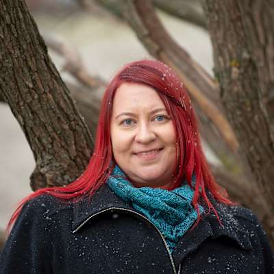 Kirjailija Hanna Lantto seisoo lumisateessa,  vanhan pajun oksien edessä, yllään tumma takki ja sininen huivi. Hänen hiuksensa ovat punaiset.