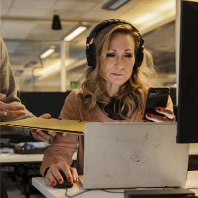 Nainen istuu työpöydän ääressä tuijottaen tietokoneen näyttöä. Hänellä on kuulokkeet korvilla, kädessään älypuhelin ja vieressä oleva henkilö ojentaa hänelle keltaista kansiota.