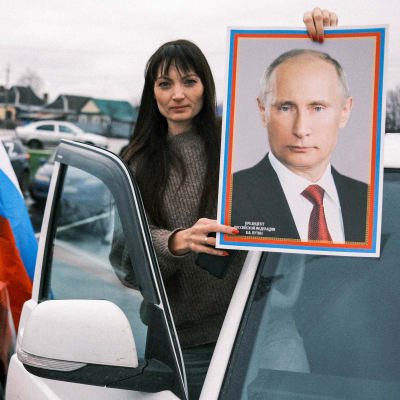 Nainen seisoo autojen välissä pidellen Putinin isoa valokuvaa, auton ikkunaan maalattu Z-kirjain.