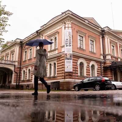 En människa promenerar på trottoaren utanför Alexandersteatern i Helsingfors. Hen har ett blått paraply.