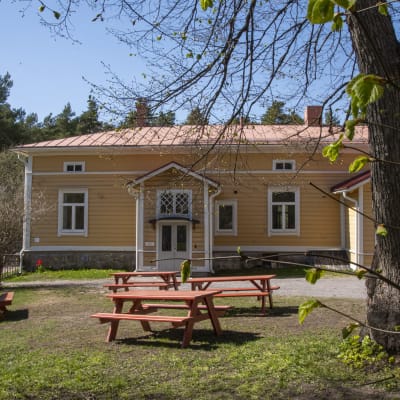Några bord och bänkar står i en trädgård vid ett gammalt gult hus. Till vänster syns en lekpark med staket omkring.