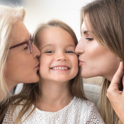 Liten leende flicka får pussar på kinden av sin mamma och mormor