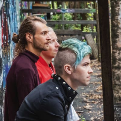 Tre män sitter på en bänk och lyssnar uppmärksamt, omgärdade av träd, i bakgrunden graffitimålning.