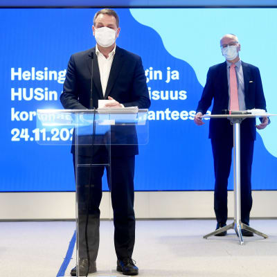 Två män står vid sina podium och håller presskonferens. 