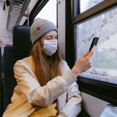 Kvinna i grå mössa och beige jacka och munskydd åker tåg, tittar på sin mobiltelefon.