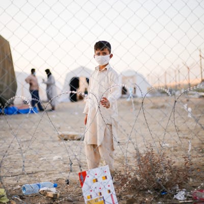En liten pojke i munskydd tittar in i kameran genom stängsel på gränsen mellan Afghanistan och Iran. I bakgrunden syns tält och två män som pratar med varandra.