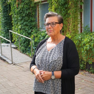 Kvinna med kort hår och glasögon står framför en gul byggnad.