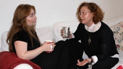 Vivi och Heidi sitter i soffan med kaffekoppar i handen och diskuterar.