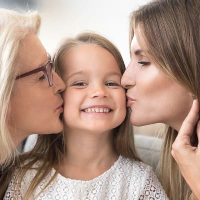 Liten leende flicka får pussar på kinden av sin mamma och mormor