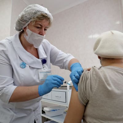 En kvinna i vit basker vaccineras av en kvinna klädd i vit jacka, hårskydd, munskydd samt blå plasthandskar.