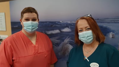 Två hälsovårdare med munskydd framför en tavla med vinterlandskap, Camilla Rautanen i röd sköterskedräkt och Carola Silander-Lehtinen i blå sköterskedräkt.