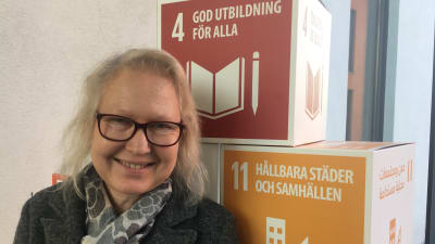 Sinikka Suomalainen, en dam med glasögon, står framför två lådor. På den ena står texten God utbildning för alla. På den andra finns texten Hållbara städer och samhällen. Hon är utbildningsplanerare vid Centret för livslångt lärande vid Åbo Akademi.
