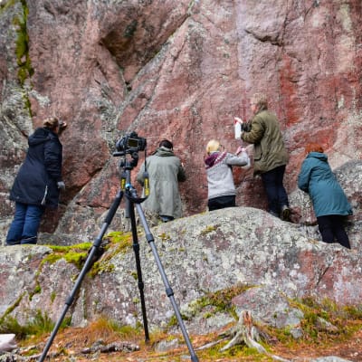 Viisi ihmistä puhdistaa muinaista kalliomaalausta korkean, punasävyisen kallion juurella.