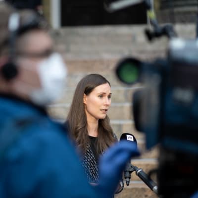 Sanna Marin ger intervjuer på Ständerhusets trappor. I förgrunden ses en kameraman som har på sig ett munskydd.