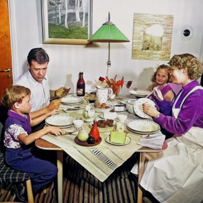 familj från 1950 talet som sitter vid ett köksbord och äter middag. Familjen ser glad och stämningen är trivsam. Modern har ett förkläde på sig. Man ser att det är hon som fixat middagen. 