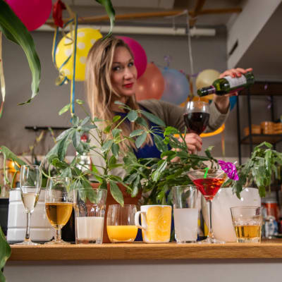 Keittiötasolla on rivissä erilaisia juomia juomalaseissa. Taustalla nainen kaataa punaviiniä lasiin. Kuvassa on myös useita viherkasveja.