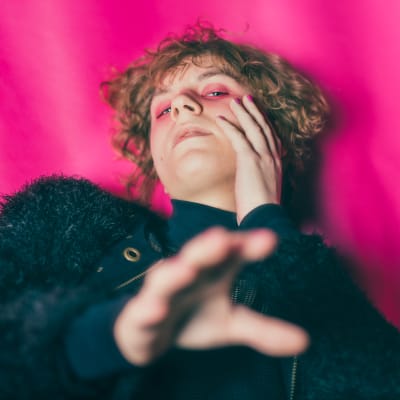 Knife Girl makaa pinkin taustan päällä ja kurkottaa kohti kameraa.