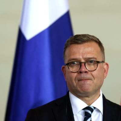 Petteri Orpo katsoo ohi kamerasta huulet tiiviisti yhdessä. Taustalla on Suomen lippu. Orpolla on pyöreät silmälasit, puku ja siniraidallinen kravatti.