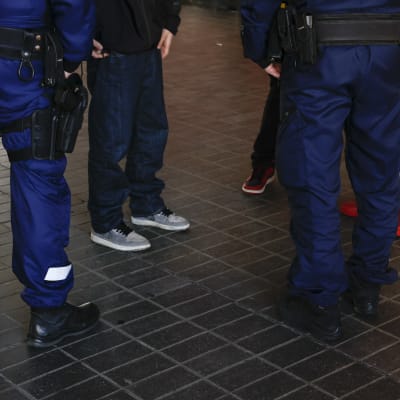 På bilden syns flera personers ben. På basis av kläderna kan man avgöra att det är två poliser som pratar med tre ungdomar.