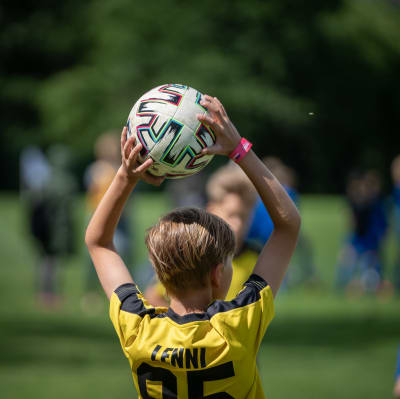 Ett barn med kort hår och fotbollströja håller en fotboll över huvudet.