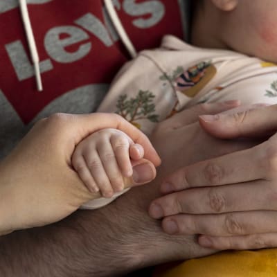 En vuxen håller en baby i handen. Två håriga armar sträcker sig runt babyn som sitter i famnen.