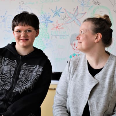 En ung person iklädd svart tröja och hens lärare iklädd grå tröja sitter framför en skoltavla med blommor ritade på.