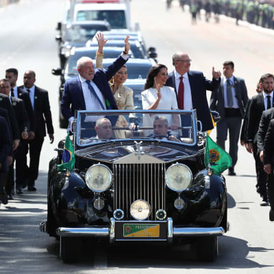 Luiz Inácio Lula da Silva och Geraldo Alckmin står och vinkar i en öppen bil tillsammans med sina fruar. På ömse sidor om bilen springer säkerhetsvakter.