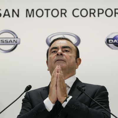 Carlos Ghosn står med knäppta händer framför en Nissan-skylt.