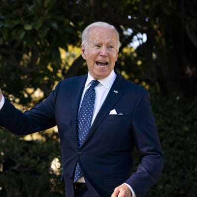Joe Biden tittar åt sidan och pratar. Han är fotograferad utomhus och är klädd i en mörkblå kostym.