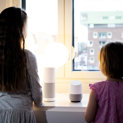 Två flickor i ca 6 och 9 årsåldern står vid ett fönster med ryggen till kameran och har två smarthögtalare framför sig som de talar till