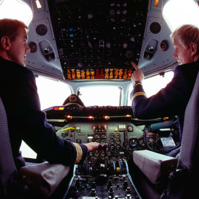 Två piloter sitter i ett flygplan vid styrpanelen. De tittar på varandra medan de sitter på varsin stol.