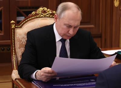 Vladimir Putin sitter vid ett skrivbord och tittar ner på ett stort papper som han håller i med båda händerna.