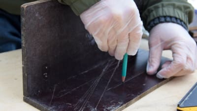 Två skivor i vattenfast faner som spikats ihop till en vinkel. En hand ritar ut markeringar på skivan med en blyertspenna.