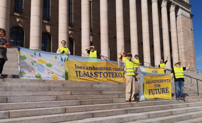 Keltaisiin liiveihin pukeutuneita mielenosoittajia eduskuntatalon portailla, pitelevät kylttejä joissa lukee "Ilmastovanhemmat vaativat ilmastotekoja".