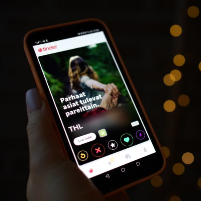 En hand som håller i en mobiltelefon. Mobilskärmen är upplåst med dejtingappen Tinder aktiverad, och på den syns Institutet för hälsa och välfärds reklambild för unga om att ta coronavaccin.