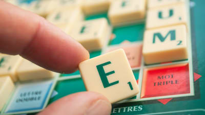 En hand håller upp bokstaven E på ett Scrabble-bräde.