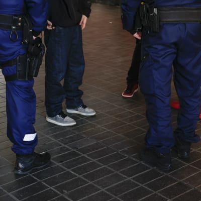 På bilden syns flera personers ben. På basis av kläderna kan man avgöra att det är två poliser som pratar med tre ungdomar.