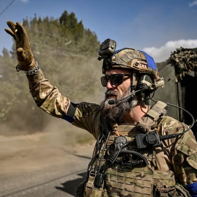En soldat fotograferad från sidan medan han vinkar. Han är klädd i full utrustning och har solglasögon på sig. Det är soligt och himlen är blå. I bakgrunden syns ett militärfordon.