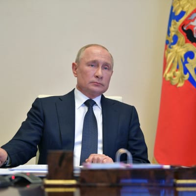 Man i blå kostym med slips ser in i kameran med neutral min. Bredvid honom står Rysslands flagga. Mannen är Rysslands president Vladimir Putin.